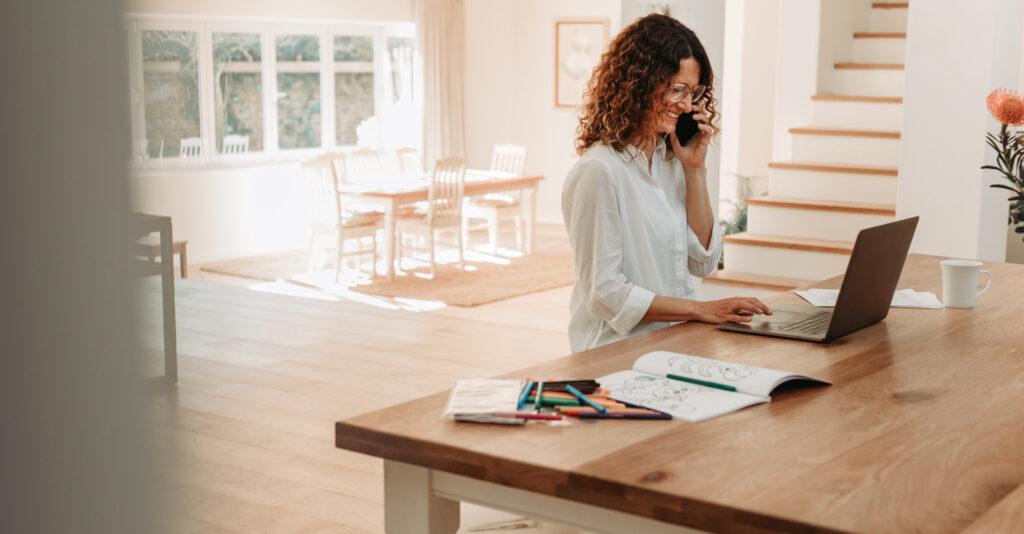 business owner standing standing at dining table with her laptop. She is on a phone call and smiling. A children's coloring book and pencils are scattered near her computer.