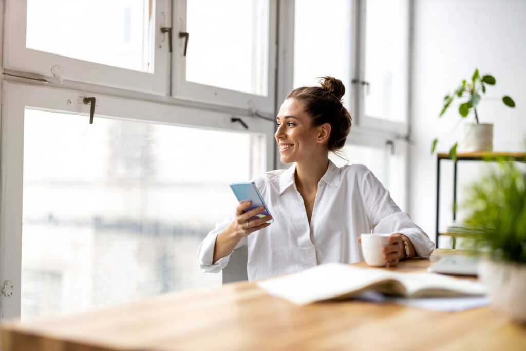 Young business owner seated at desk in an airy office, holding a cup of coffee and her cell phone. She is looking out the window and smiling in a relaxed way.