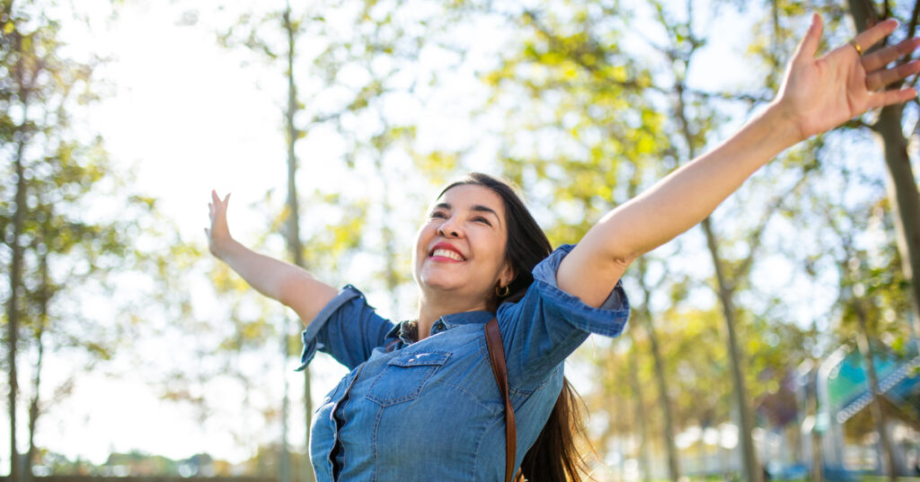 smiling woman in park. She's stretching her arms upward in the sunshine and smiling broadly.