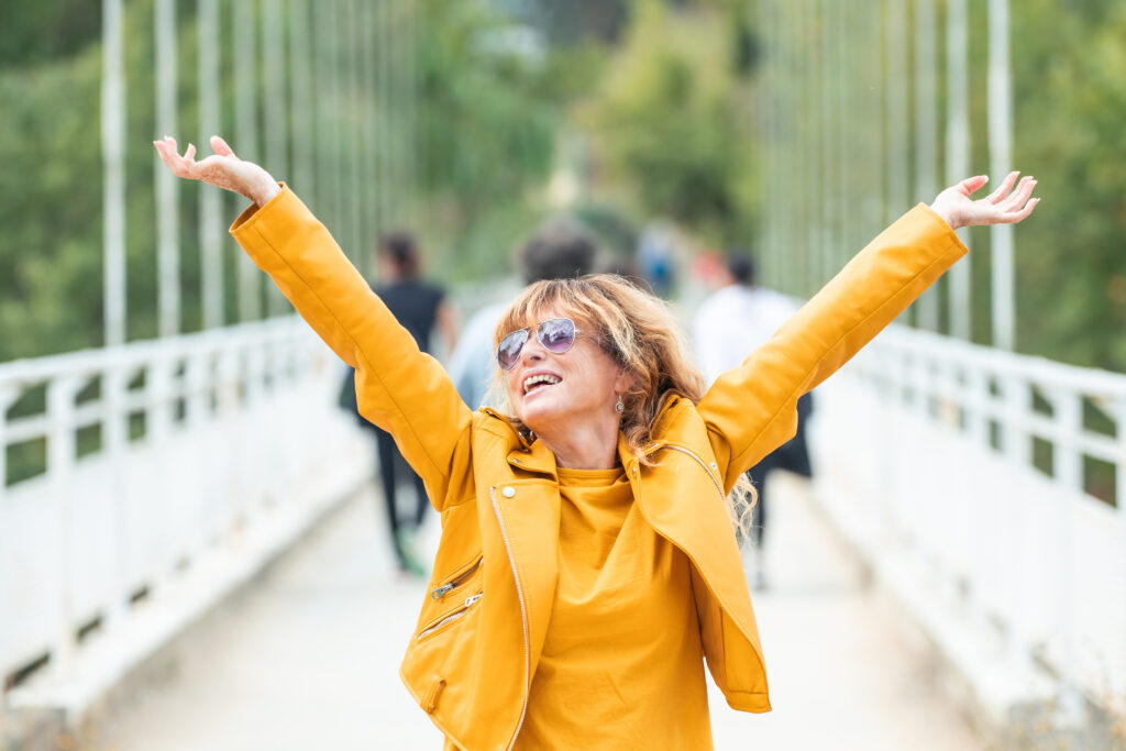 Photo of middle aged woman wearing sunglasses and a bright yellow moto jacket. She is stretching both arms upward with energy and smiling with joy.
